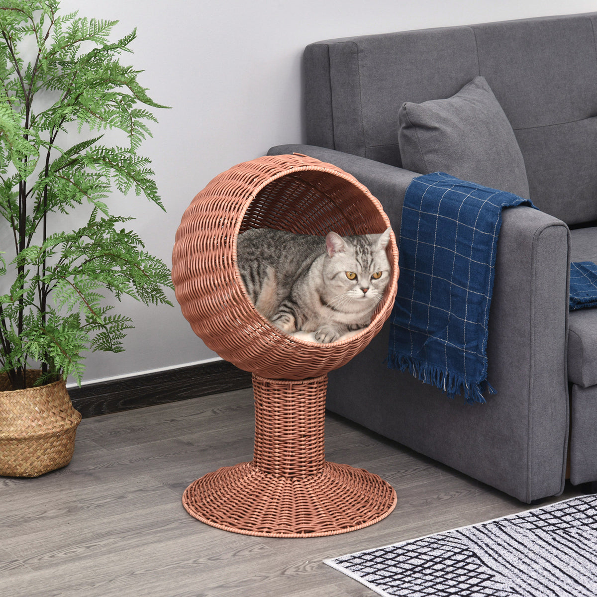 Elevated wicker cat basket bed with swivel pod beside a gray sofa and houseplant